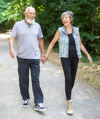 Ein Seniorenpaar geht lachend Hand in Hand auf einem Waldweg.
