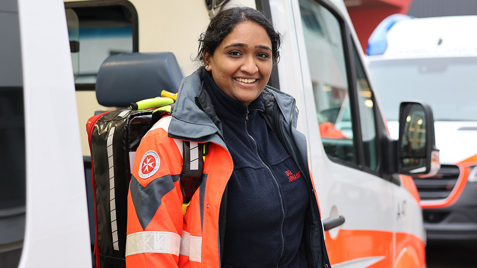 Myurri steigt lächelnd aus einem Rettungswagen. Sie trägt die orangene Johanniter-Uniform