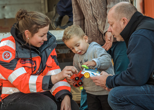 Ein Junge spielt mit seinen neuen Spielzeugautos. Neben ihm knien sein Vater und eine Johanniterin.