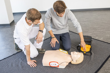 Ein Mann betätigt im Kurs den Defibrillator an einer Puppe. Neben ihm sitzt der Kursleiter und schaut ihm zu.
