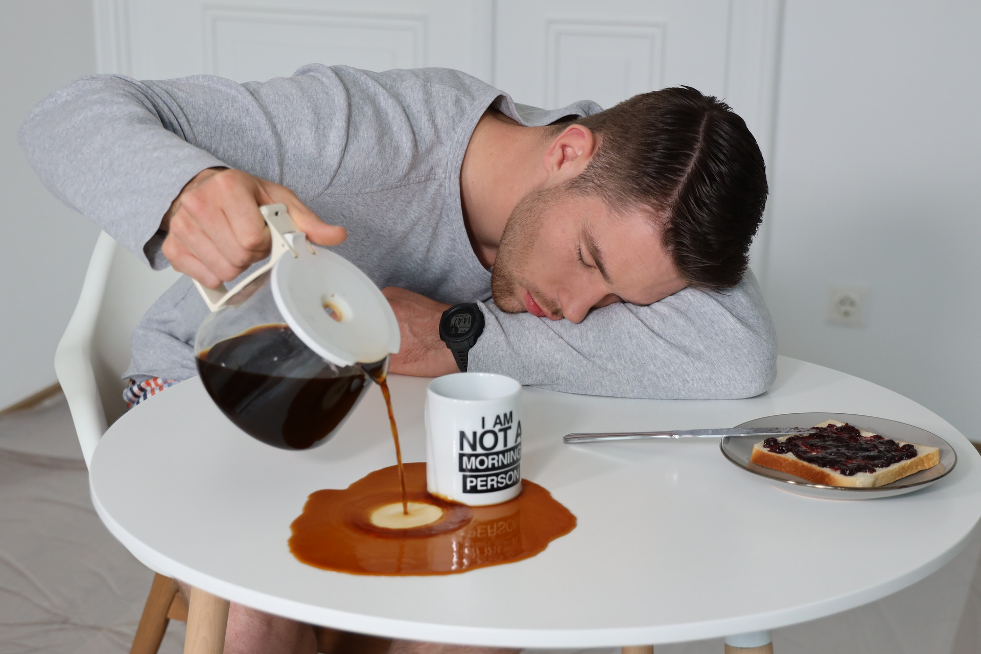 Ein Mann, Johannes, sitzt beim Frühstück. Er ist so müde, dass er den Kaffee aus der Kanne beim Einschenken daneben schüttet.