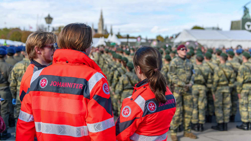 Drei Sanitäter:innen beobachten die Feierlichkeiten beim Nationalfeiertag. Im Hintergrund stehen Soldaten für die Angelobung.