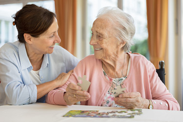Eine Pflegerin sitzt neben einer älteren Dame, die ein Puzzle löst.
