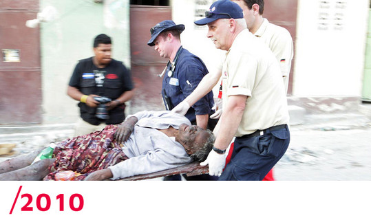 2010: Drei Helfer transportieren einen verletzten Mann auf einer Trage ab. Im Hintergrund steht ein Reporter.