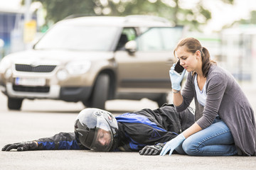 Eine Frau sitzt neben einem verunfallten Motorradfahrer und ruft mit dem Telefon nach Hilfe.