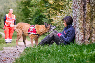 Ein Rettungshund hat eine Vermisste gefunden und bellt um auf sich aufmerksam zu machen. Im Hintergrund steht eine Johanniterin.