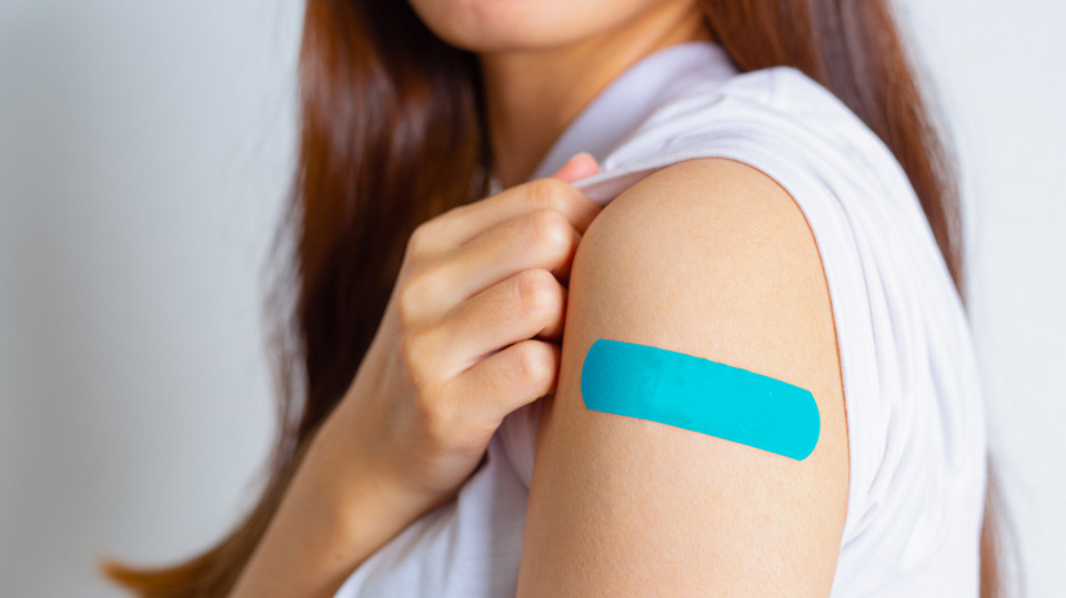 Eine Frau zeigt ihren Oberarm, wo ein blaues Pflaster vom Impfen ist.