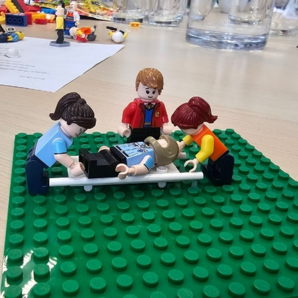 Im Bild sieht man Legomännchen, die als Sanitäter:innen einen Patienten auf der Trage versorgen.