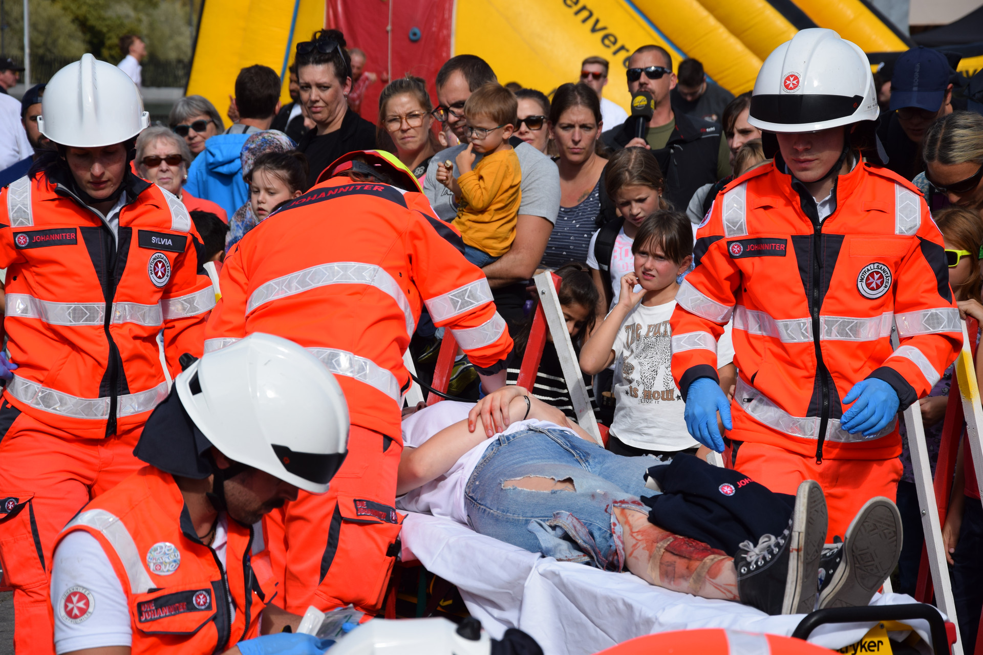 Vier Johanniter versorgen eine Patientin. Im Hintergrund steht das Publikum der Leistungsschau.