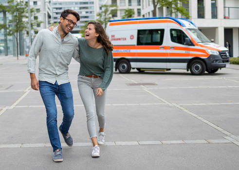 Zwei Ehrenamtliche gehen lachend nebeneinander. Im Hintergrund steht ein Rettungswagen.