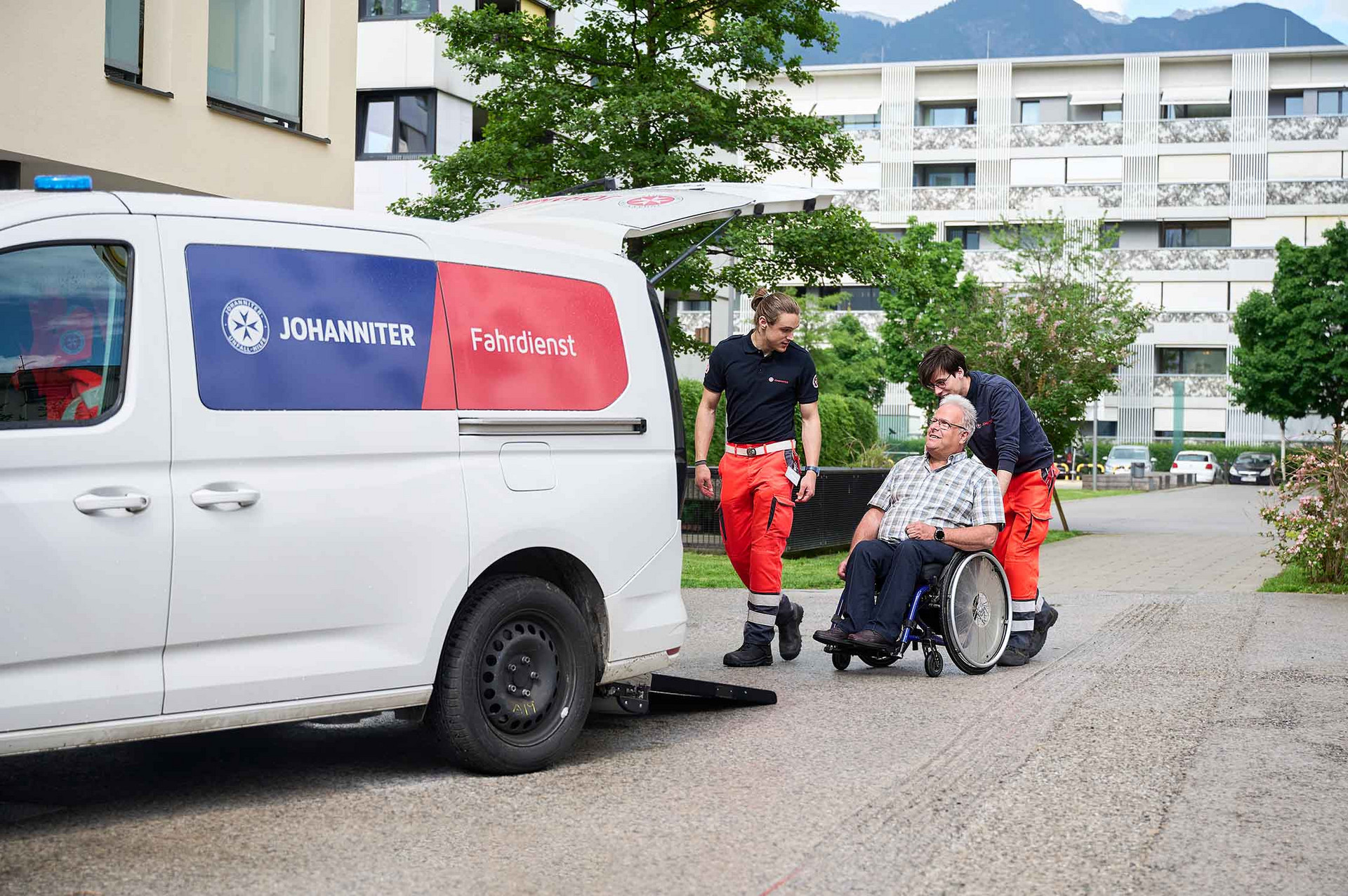 Zwei Johanniter vom Fahrdienst schieben einen Klienten im Rollstuhl zum Auto.