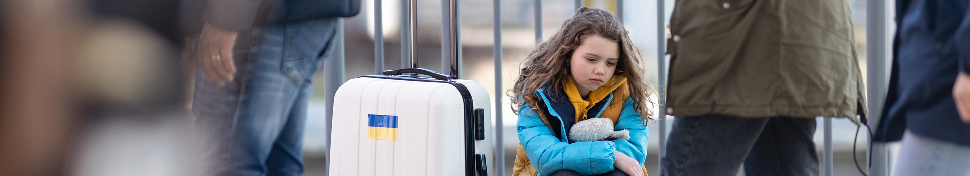 Ein trauriges Mädchen sitzt mit mehreren Gepäckstücken am Bahnhof. Am Koffer ist ein Ukraine-Sticker.