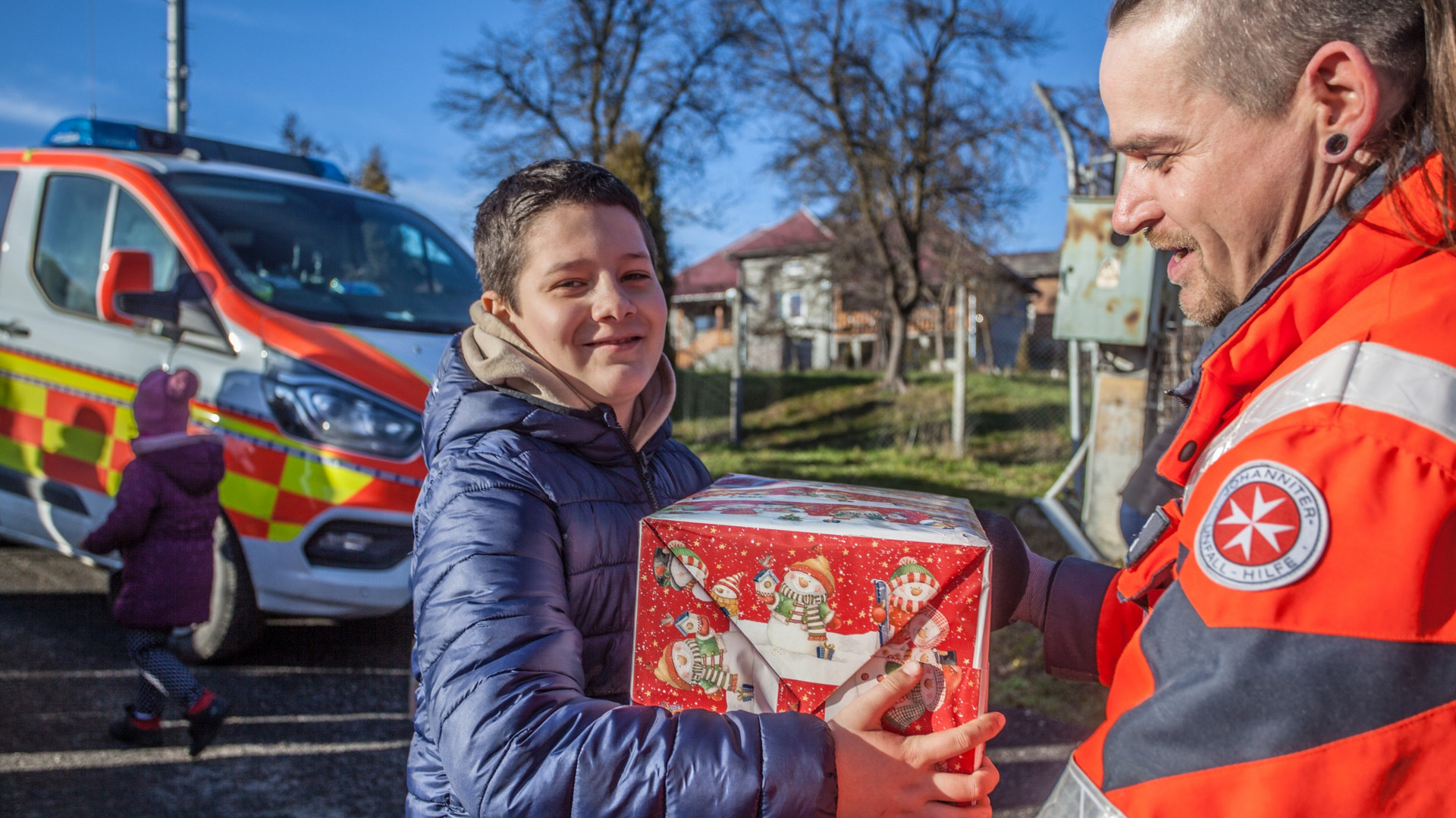 ein Helfer in Johanniter Uniform überreicht einem lächelnden Kind ein Paket