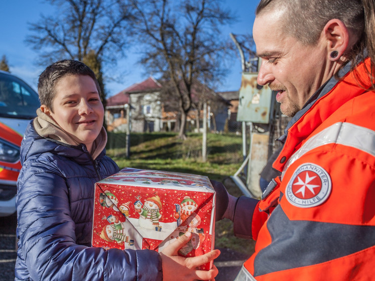 ein Helfer in Johanniter Uniform überreicht einem lächelnden Kind ein Paket