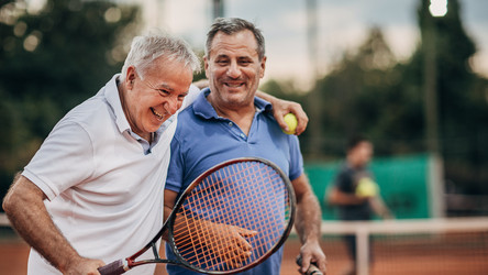 Zwei ältere Männer sind auf einem Tennisplatz und haben Tennisschläger und einen Tennisball in der Hand.
