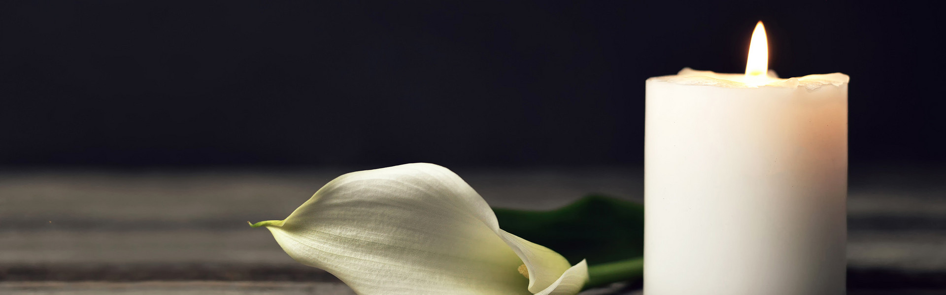 Eine Kalla und eine brennende weiße Kerze vor schwarzem Hintergrund.