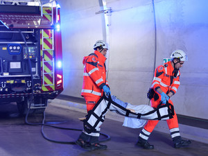 zwei Sanitäter tragen eine Übungspuppe, im Hintergrund sieht man ein Feuerwehrauto mit Blaulicht