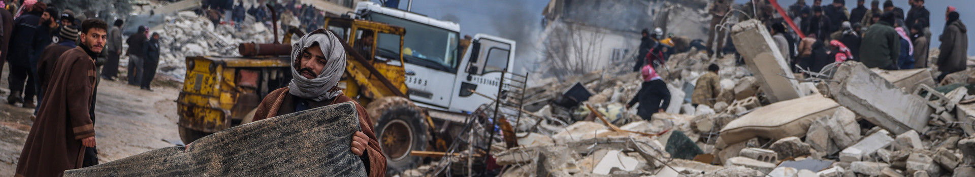 Mann trägt eine schwarze Matraze, im Hintergrund sind zerstörrte Gebäude zu sehen
