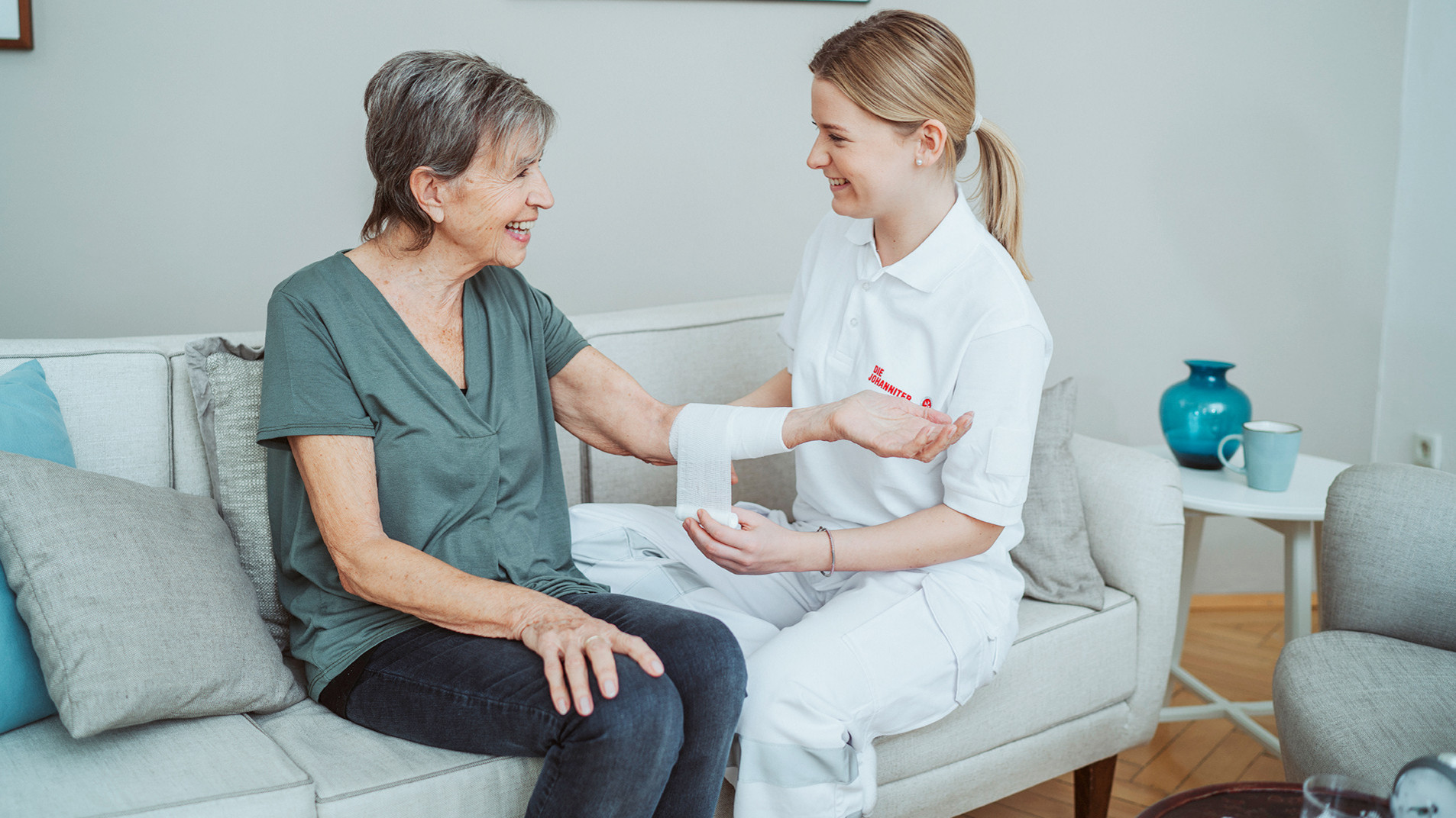 Eine Pflegerin sitzt mit der Patientin auf dem Sofa und legt ihr am Unterarm einen Verband an.