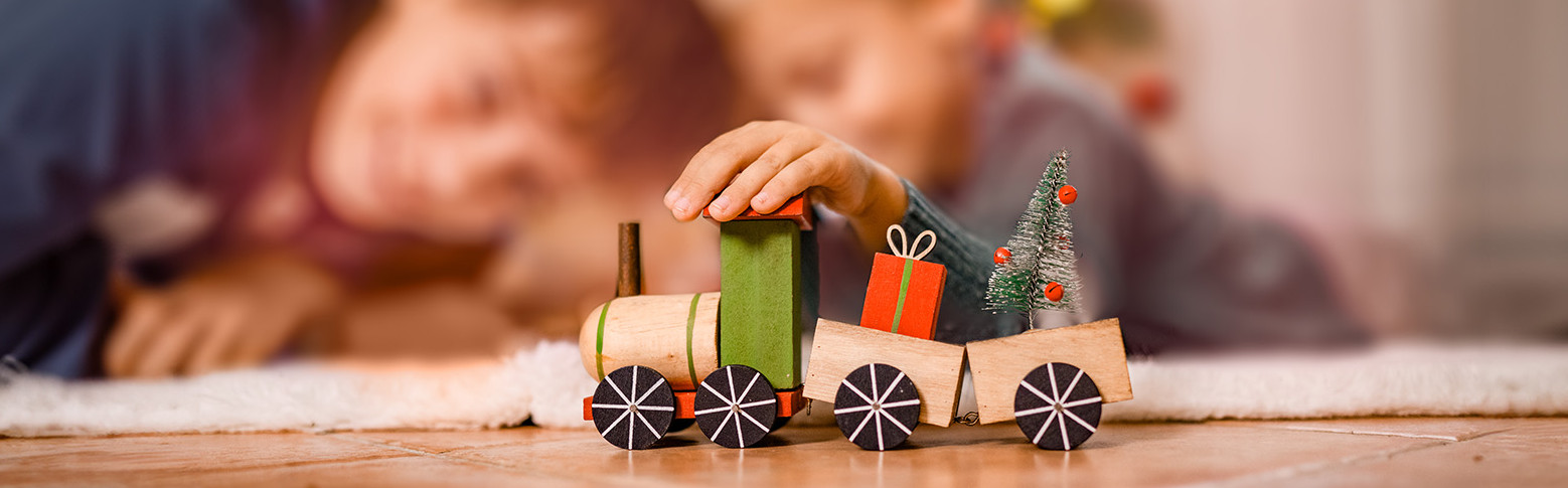 Zwei Kinder spielen an Weihnachten mit einem Spielzeugzug vor dem Christbaum.