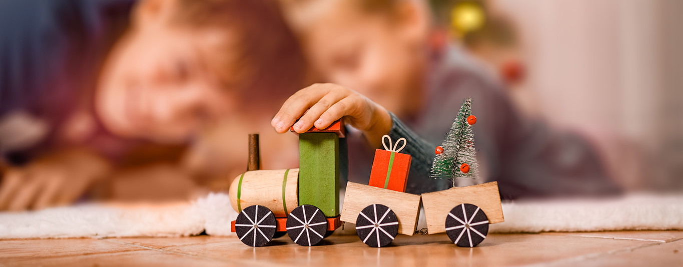 Zwei Kinder spielen an Weihnachten mit einem Spielzeugzug vor dem Christbaum.