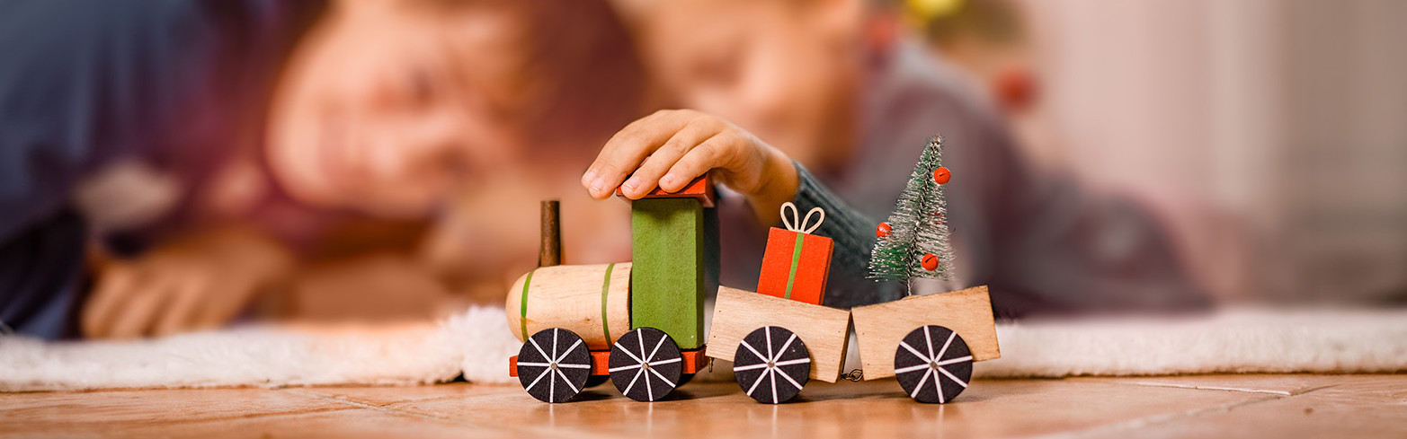 Zwei Kinder spielen an Weihnachten mit einem Spielzeugzug vor dem Christbaum.