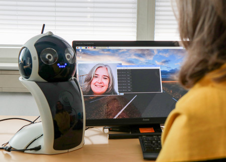Eine Frau sitzt vor einem Computer. Daneben steht ein kleiner Roboter vom Projekt "RoboGen", der mit Menschen interagieren kann.