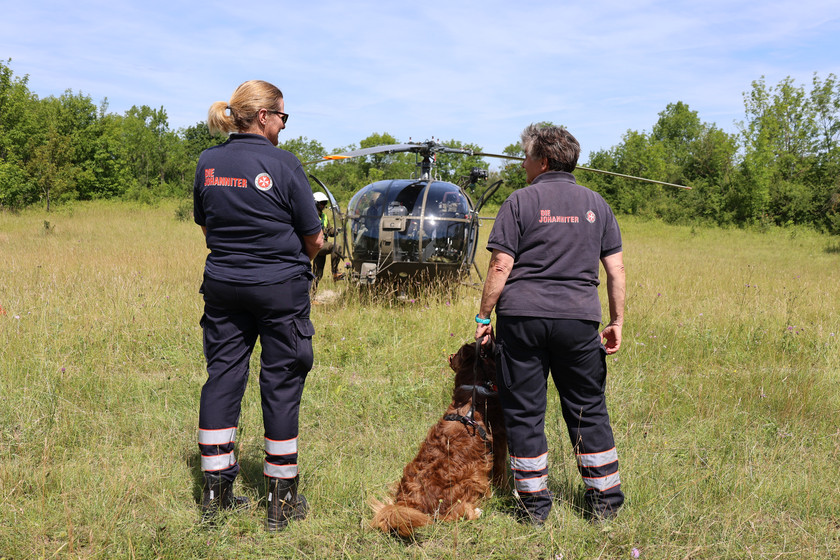 Zwei Johanniterinnnen der Rettungshundestaffel und ein Rettungshund stehen mit dem Rücken zur Kamera. Zwischen den beiden Frauen sieht man im Hintergrund einen Hubschrauber.
