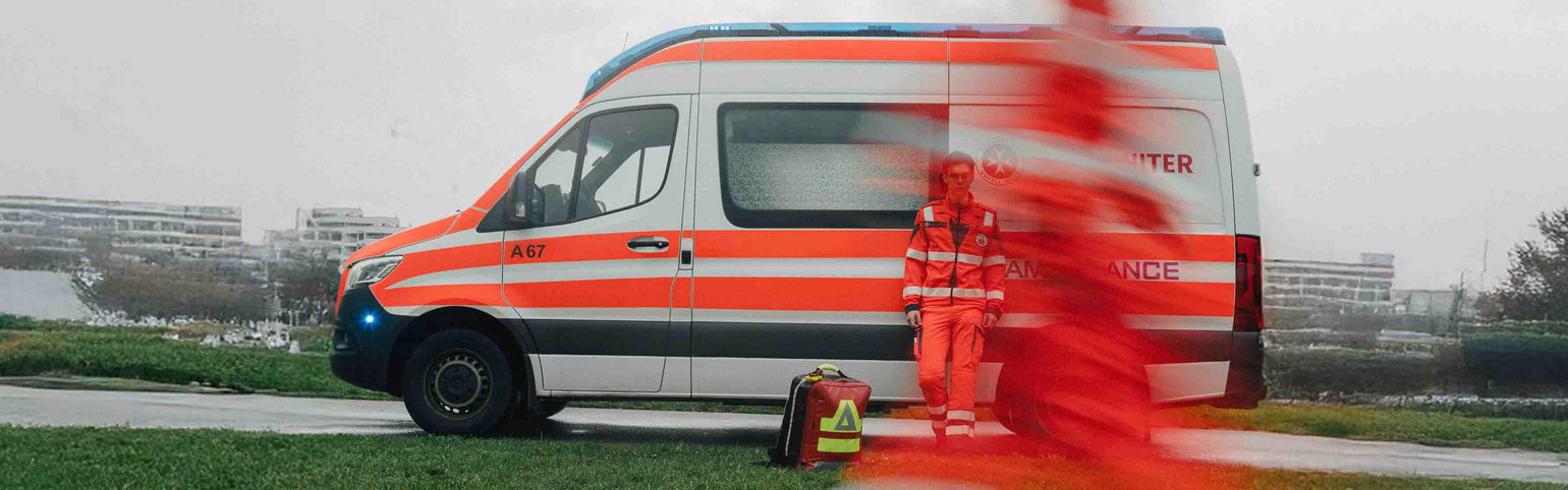 Ein Johanniter-Rettungsauto steht auf einer Straße. Daran lehnt ein Zivildiener, während ein zweiter im Vordergrund vorbeiläuft.