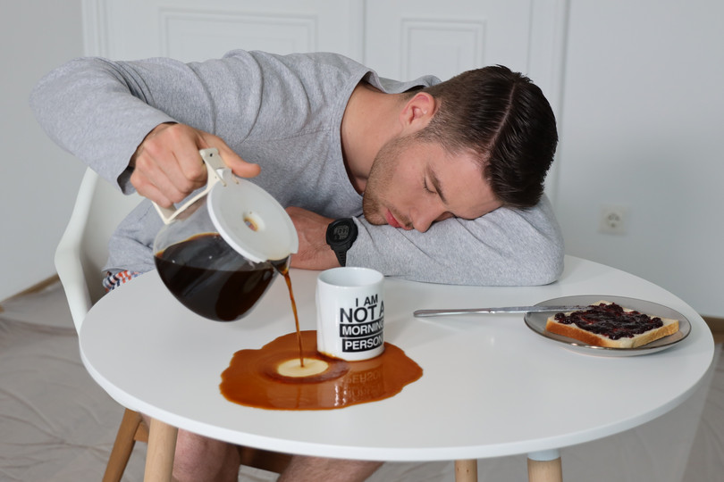 Ein Mann, Johannes, sitzt beim Frühstück. Er ist so müde, dass er den Kaffee aus der Kanne beim Einschenken daneben schüttet.
