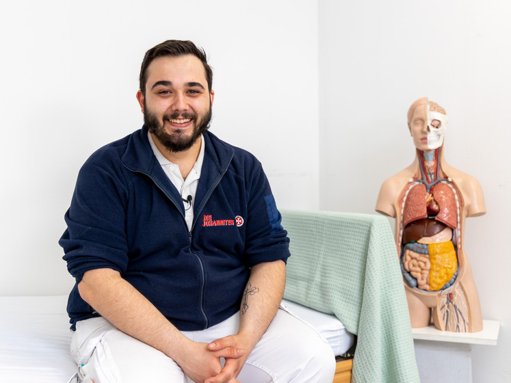 Ein Pflegeassistent, Christian, sitzt auf einem Krankenbett und lächelt in die Kamera. Im Hintergrund steht ein medizinisches Modell des Körpers.