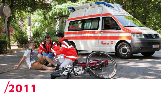 2011: Zwei Johanniter verarzten einen vom Fahrrad gestürzten Jungen. Im Hintergrund steht der Rettungswagen.