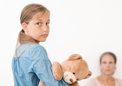 Ein Mädchen steht mit einem Teddybären in der Hand vor dem Bett ihrer Mutter, die sie pflegen muss.