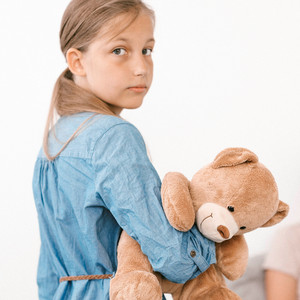 Ein Mädchen steht mit einem Teddybären in der Hand vor dem Bett ihrer Mutter, die sie pflegen muss.