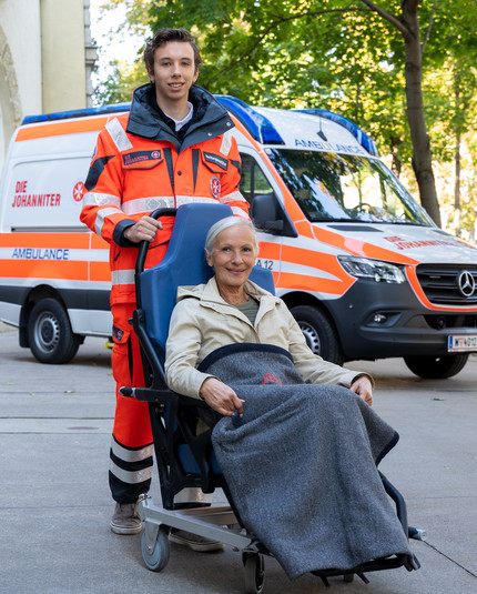 Ein Zivildienstleistender schiebt einen Tragsessel mit einer Patientin. Im Hintergrund ist ein Krankentransportwagen.