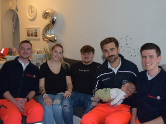 Fünf Personen sitzen auf einem Sofa und lächeln in die Kamera. Drei Personen tragen eine Saniäteruniform, einer hält Baby im Arm