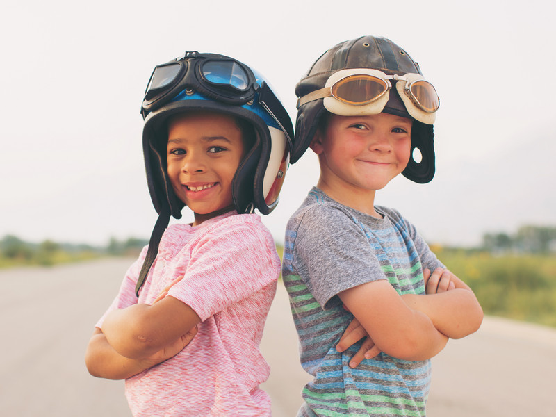 Zwei Kinder mit Fliegerhaube stehen lächelnd mit verschränkten Armen Rücken an Rücken