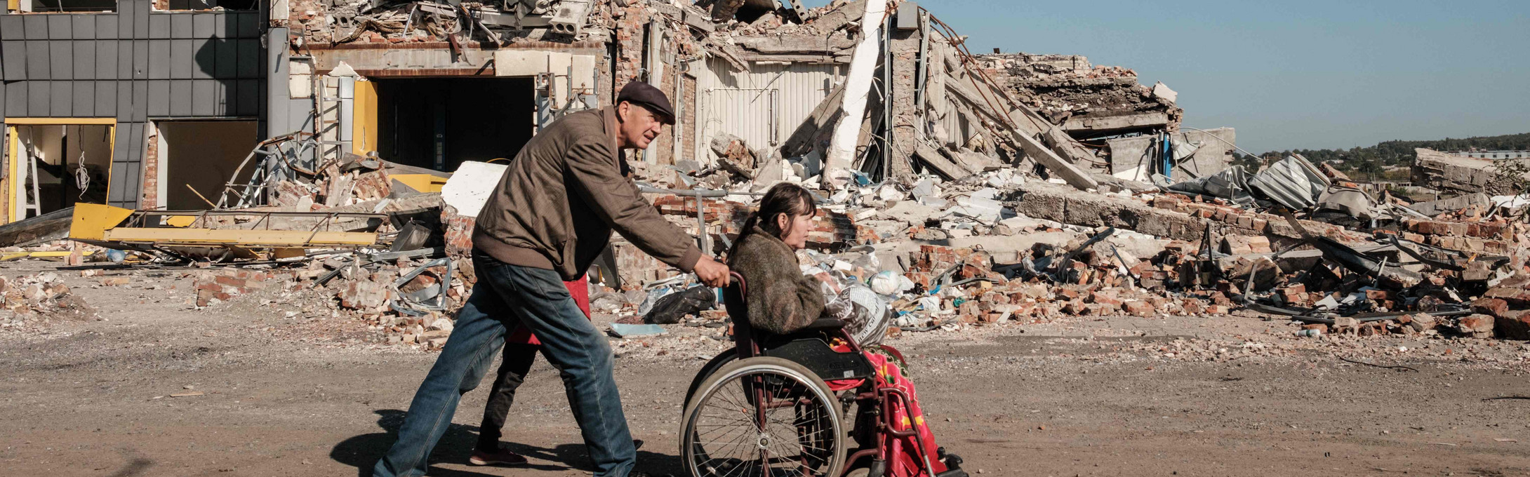 Mann schiebt Frau in Rollstuhl, im Hintergrund sind zerstörrte Häuser zu sehen