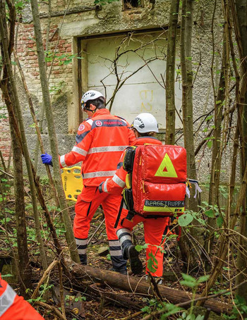 Zwei Johanniter gehen mit Rettungsrucksack und Trage im Wald zu einem eingestürzten Haus.