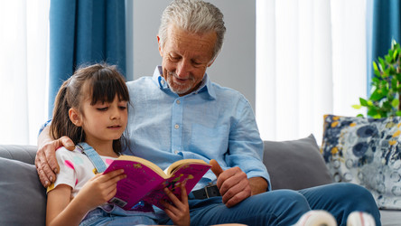 Ein Großvater sitzt mit seiner Enkelin auf der Couch und ließt mit ihr ein Buch.