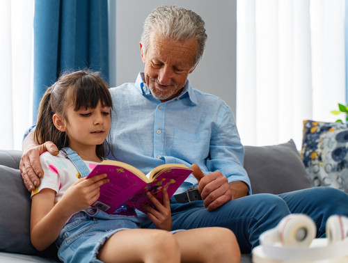 Ein Großvater sitzt mit seiner Enkelin auf der Couch und ließt mit ihr ein Buch.
