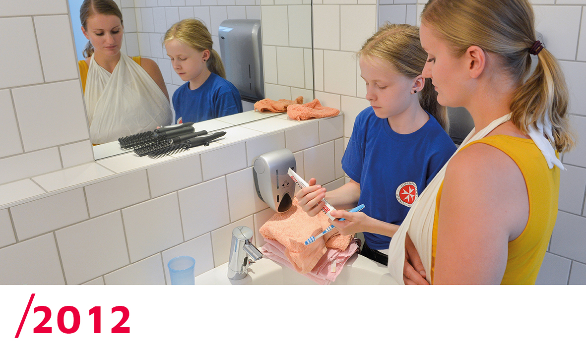 2012: Ein Mädchen steht mit ihrer Mutter, die einen Armverband hat, im Bad und gibt für sie Zahnpasta auf die Zahnbürste.