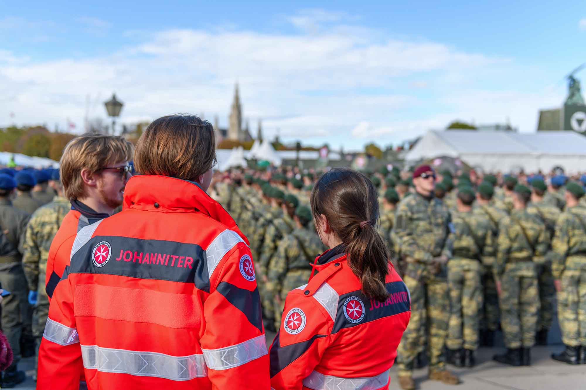 Drei Sanitäter:innen beobachten die Feierlichkeiten beim Nationalfeiertag. Im Hintergrund stehen Soldaten für die Angelobung.